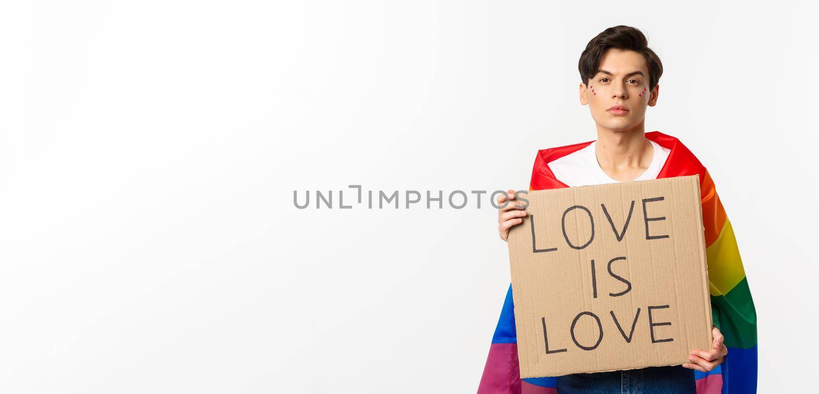 Serious and confident gay man wearing rainbow lgbt flag, holding sign for pride parade, standing over white background.