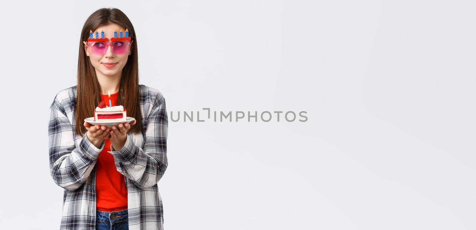 People lifestyle, holidays and celebration, emotions concept. Cute and silly birthday girl in funny glasses, look away think of wish as blowing lit candle on b-day cake, smiling excited by Benzoix