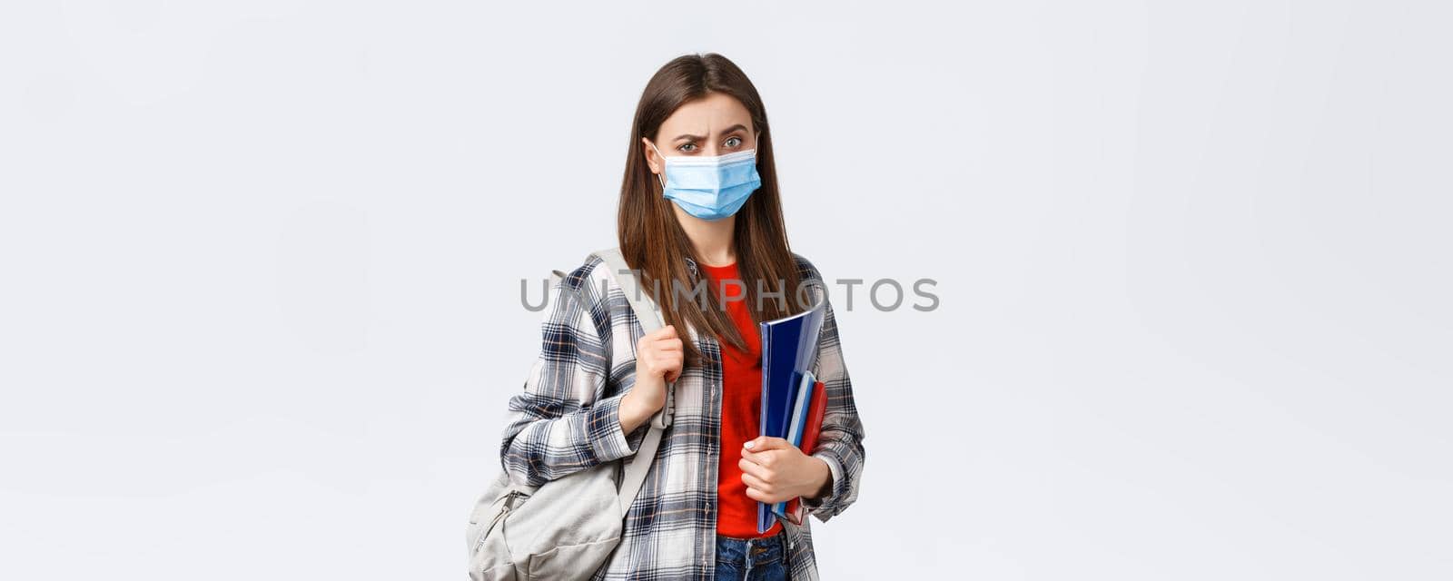 Coronavirus pandemic, covid-19 education, and back to school concept. Doubtful and skeptical female student in medical mask frowning disappointed, hold notebooks and backpack.