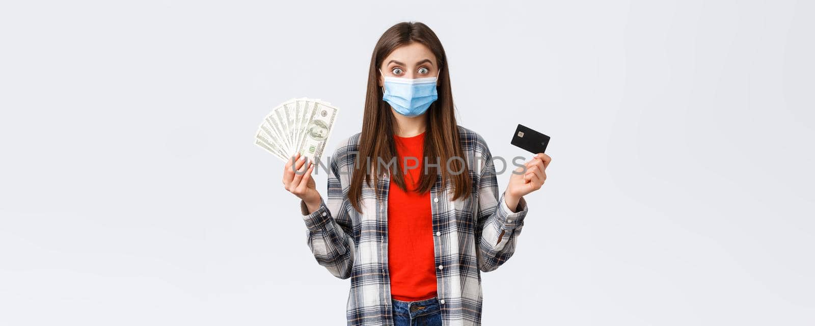 Money transfer, investment, covid-19 pandemic and working from home concept. Excited young woman earn or win lots of cash, showing dollars and credit card, wearing medical mask by Benzoix