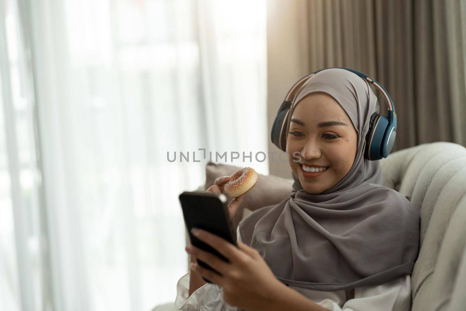 Beautiful Asian muslim woman using mobile phone while sitting on sofa, girl doing chat text message on smartphone and smiling.