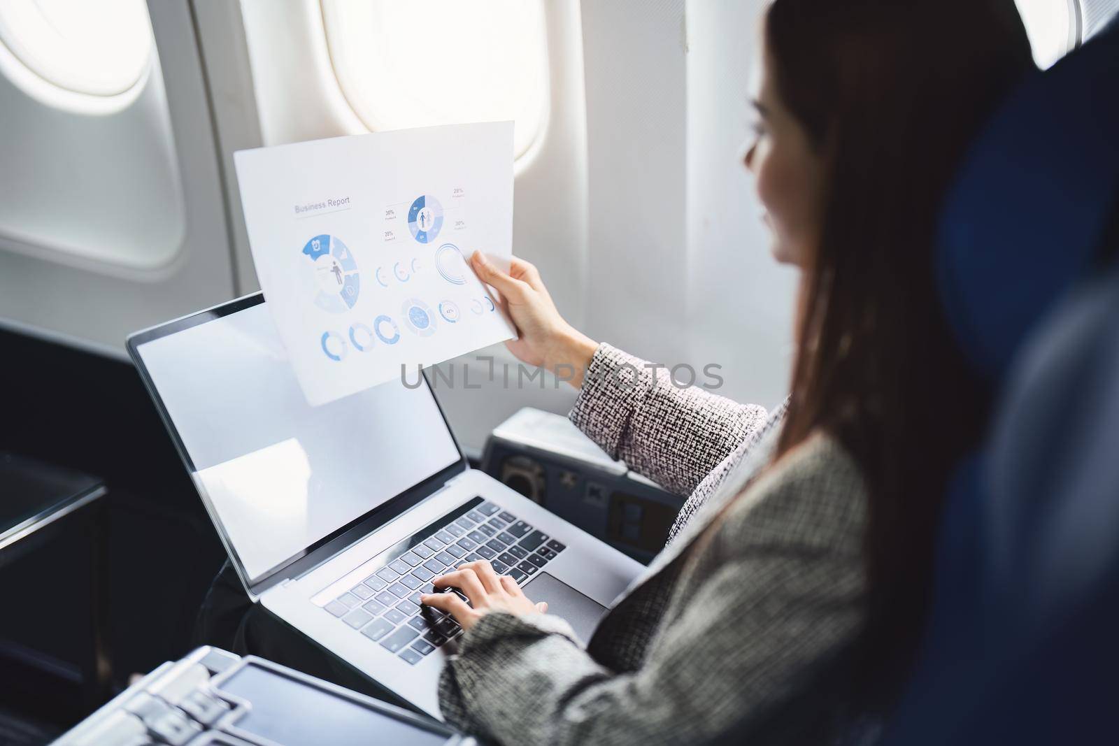 A successful asian business woman or female entrepreneur in formal suit in a plane sits in a business class seat and uses a documents with computer laptop during flight.