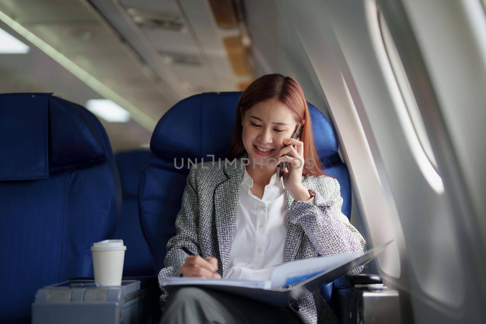 portrait of A successful asian business woman or female entrepreneur in formal suit in a plane sits in a business class seat and uses smartphone with documents for work during flight.