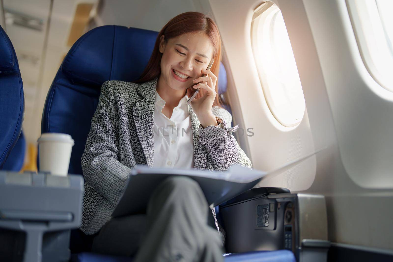 portrait of A successful asian business woman or female entrepreneur in formal suit in a plane sits in a business class seat and uses smartphone with documents for work during flight.