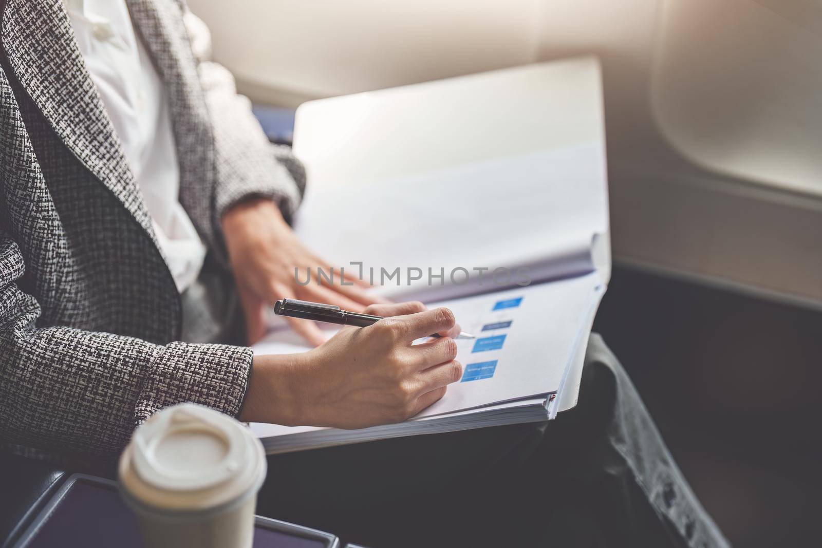 A successful asian business woman or female entrepreneur in formal suit in a plane sits in a business class seat and uses a pen with documents for work during flight.