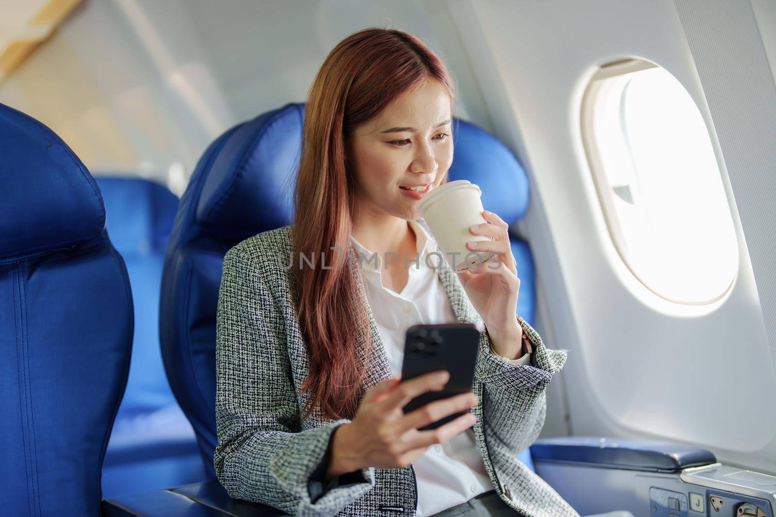 portrait of A successful asian business woman or female entrepreneur in formal suit in a plane sits in a business class seat and uses a smartphone with drink coffee during flight, relax concept.