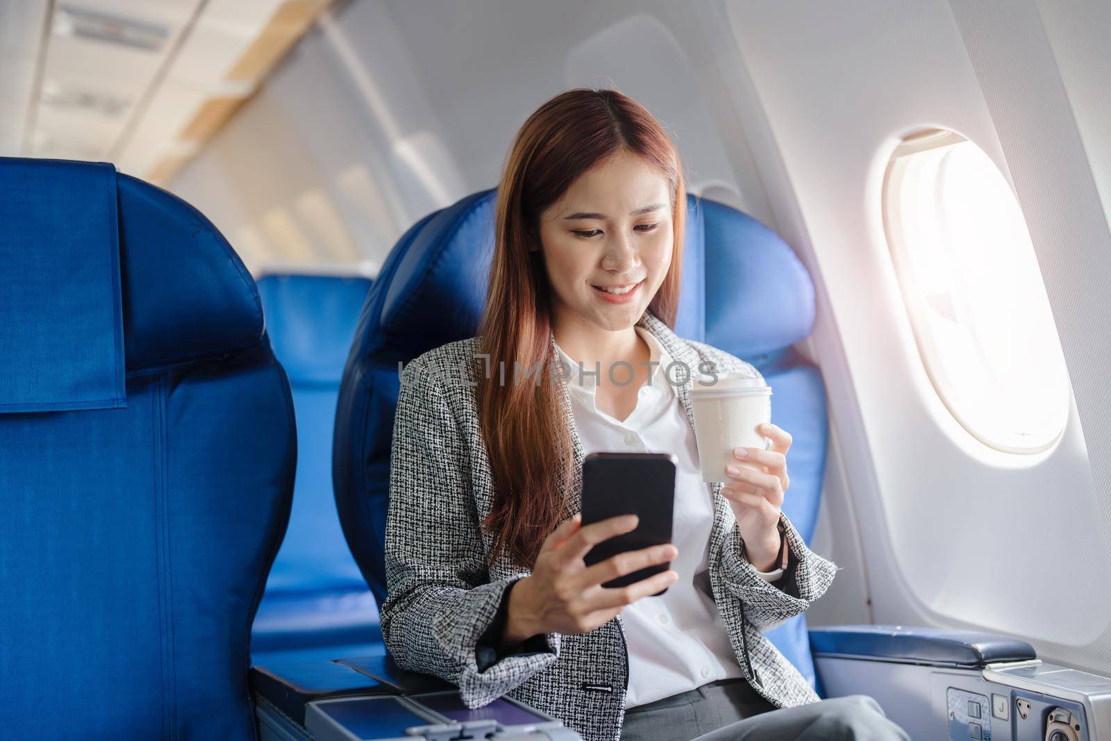 portrait of A successful asian business woman or female entrepreneur in formal suit in a plane sits in a business class seat and uses a smartphone with drink coffee during flight, relax concept.