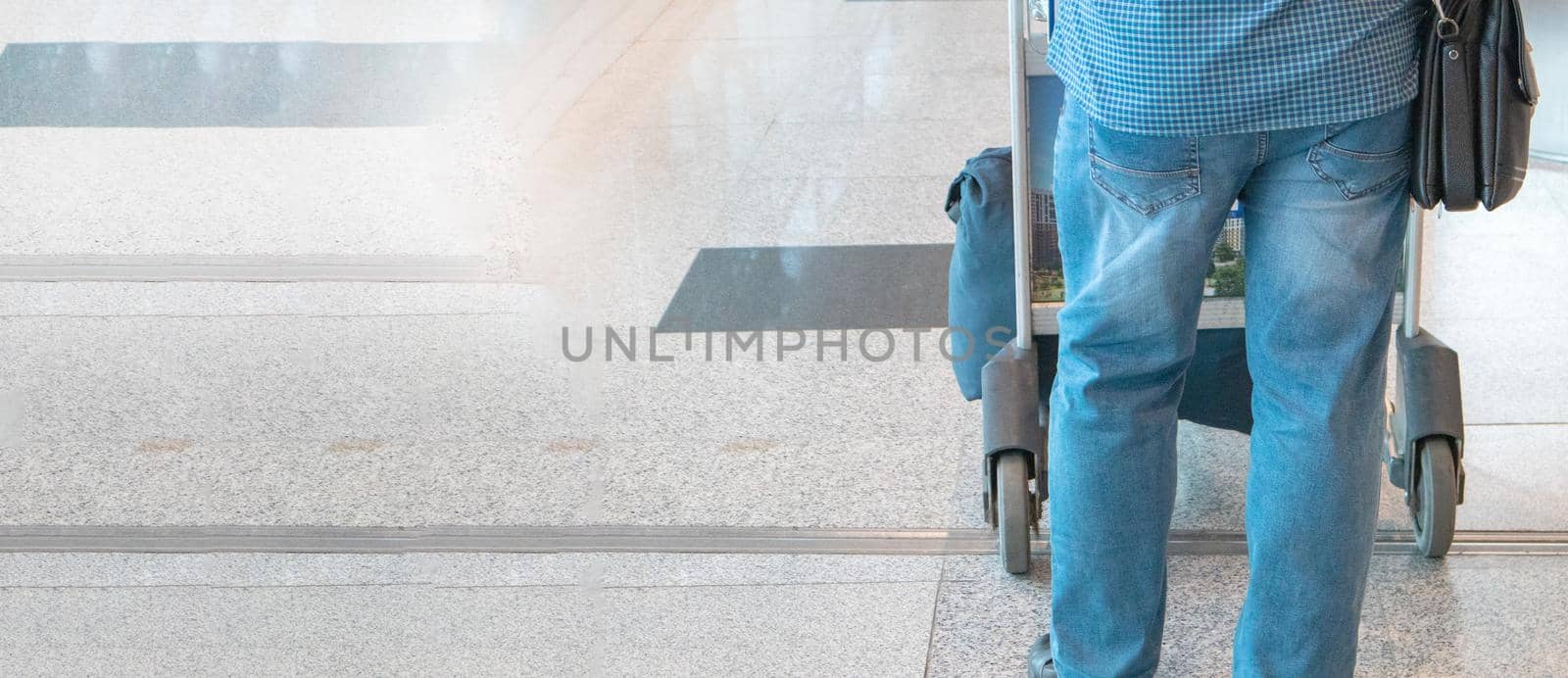 Banner a male passenger in blue jeans and a shirt, pushing a trolley with luggage at the airport, a view from the back, a copy of the space on the left.