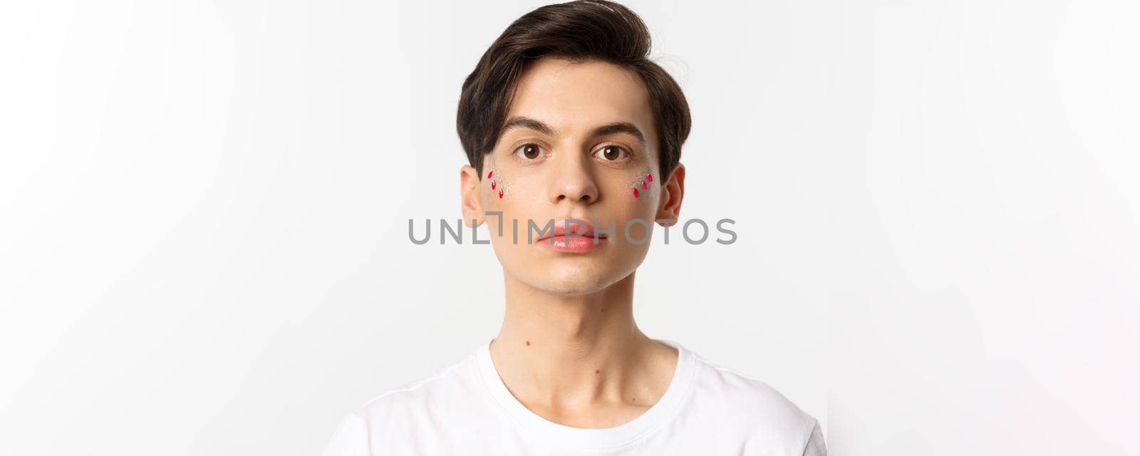 People, lgbtq and beauty concept. Close-up of beautiful androgynous man with glitter on face, looking at camera, standing over white background.