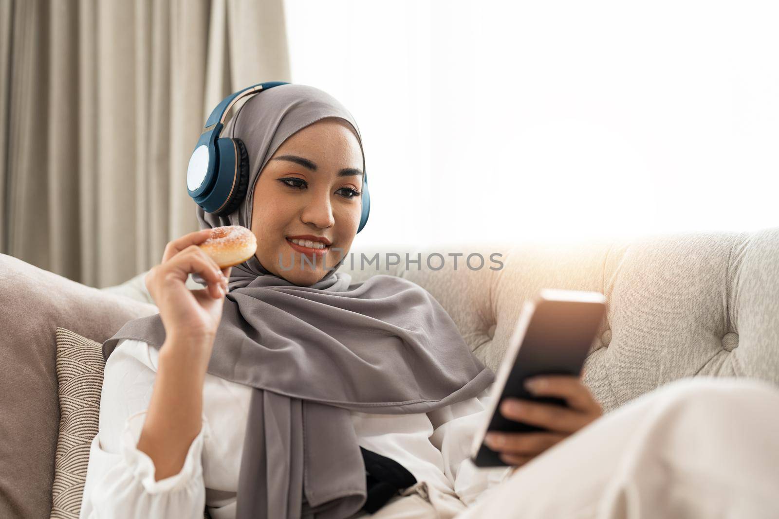 Beautiful Asian muslim woman using mobile phone while sitting on sofa, girl doing chat text message on smartphone and smiling by nateemee