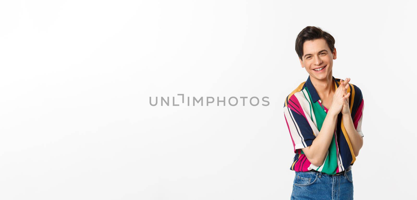 Attractive young slim man relish something good, rubbing hands and smiling satisfied, standing over white background. Copy space