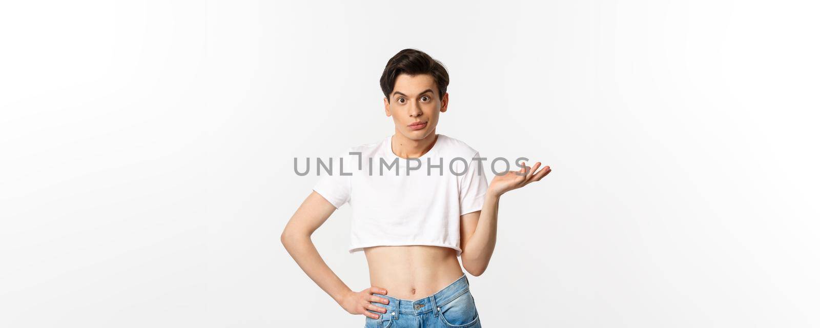 Handsome androgynous man in crop top looking confused, shrugging clueless, standing over white background.