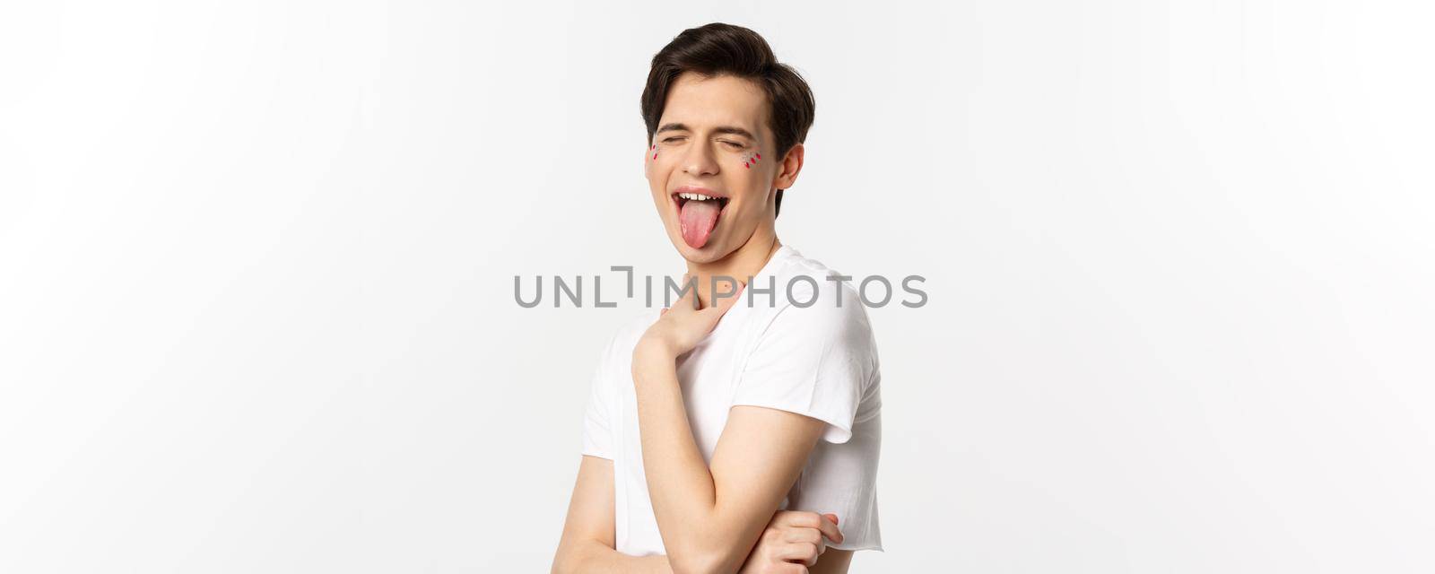 People, lgbtq and beauty concept. Close-up of carefree gay man with glitter on face, showing tongue and making funny face, standing over white background.