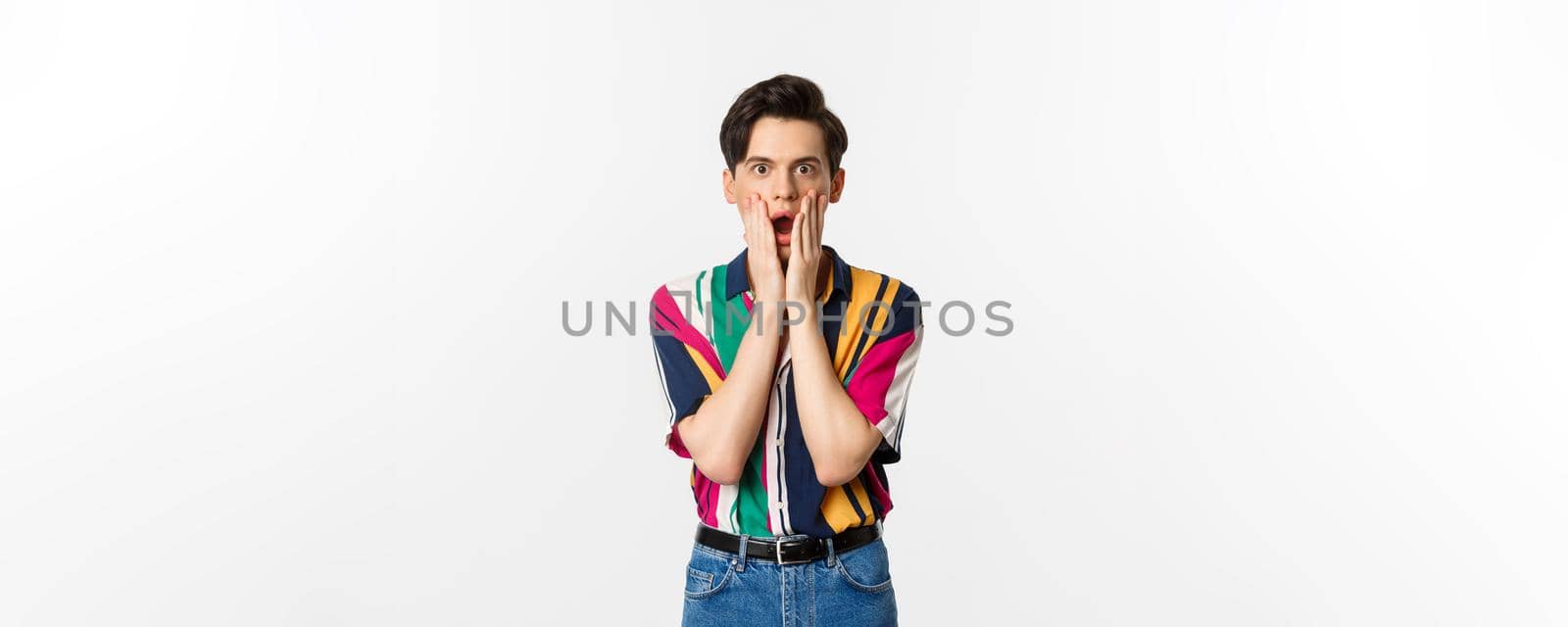 Portrait of handsome androgynous man looking shocked, gasping and holding hands on face, stare at camera amazed, standing over white background.