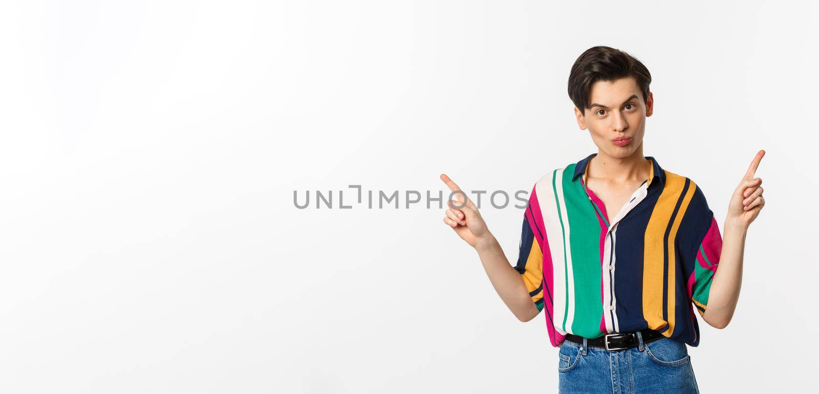Handsome gay man pointing fingers sideways, showing two variants, demonstrate promos, standing over white background.