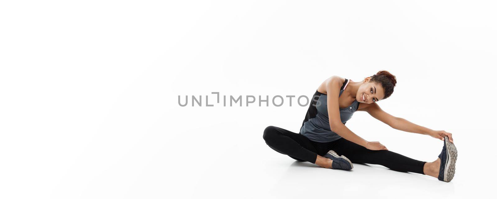 Sport, training, lifestyle and Fitness concept - portrait of beautiful happy African American woman stretching leg while sitting. Isolated on white studio background. by Benzoix