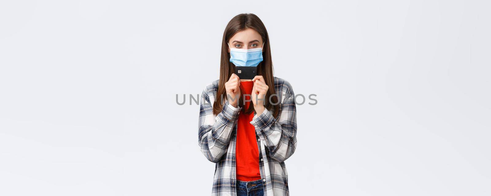 Coronavirus outbreak, working from home, online shopping and contactless payment concept. Girl in medical mask smiling sly and showing credit card, ready waste all money in internet stores.