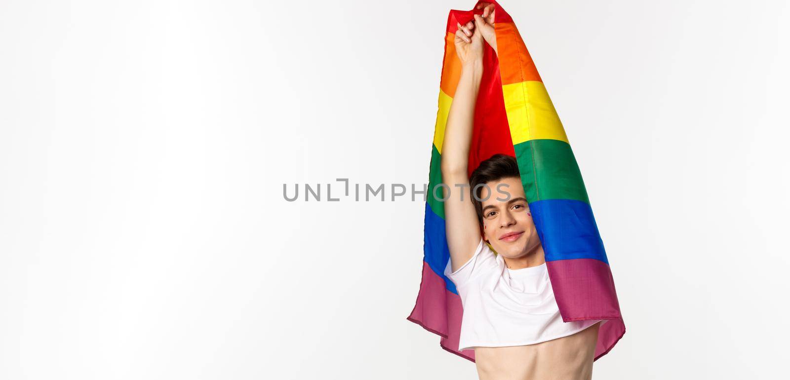 Vertical view of beautiful androgynous gay man raising rainbow flag and smiling happy, standing in crop top and jeans against white background by Benzoix