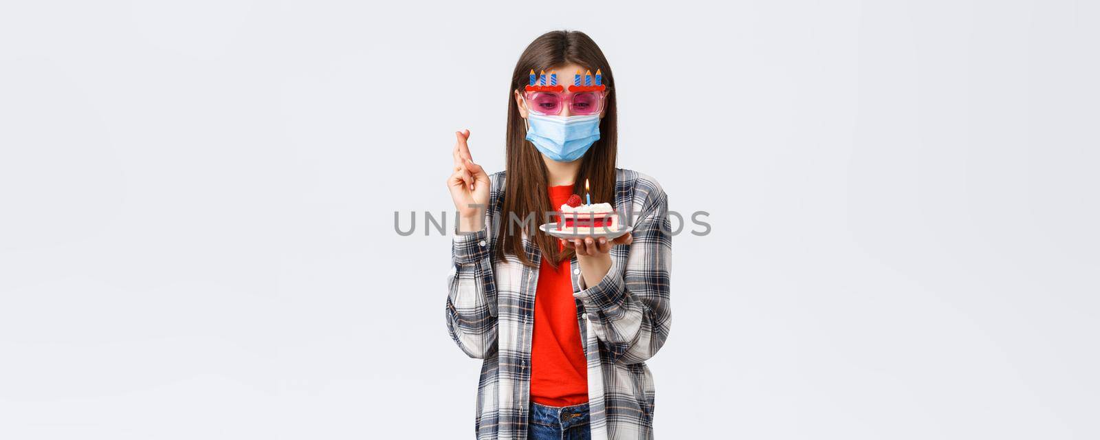 Coronavirus outbreak, lifestyle social distancing and celebration concept. Enthusiastic happy girl in glasses and medical mask celebrate birthday on quarantine, cross fingers, make wish at cake.
