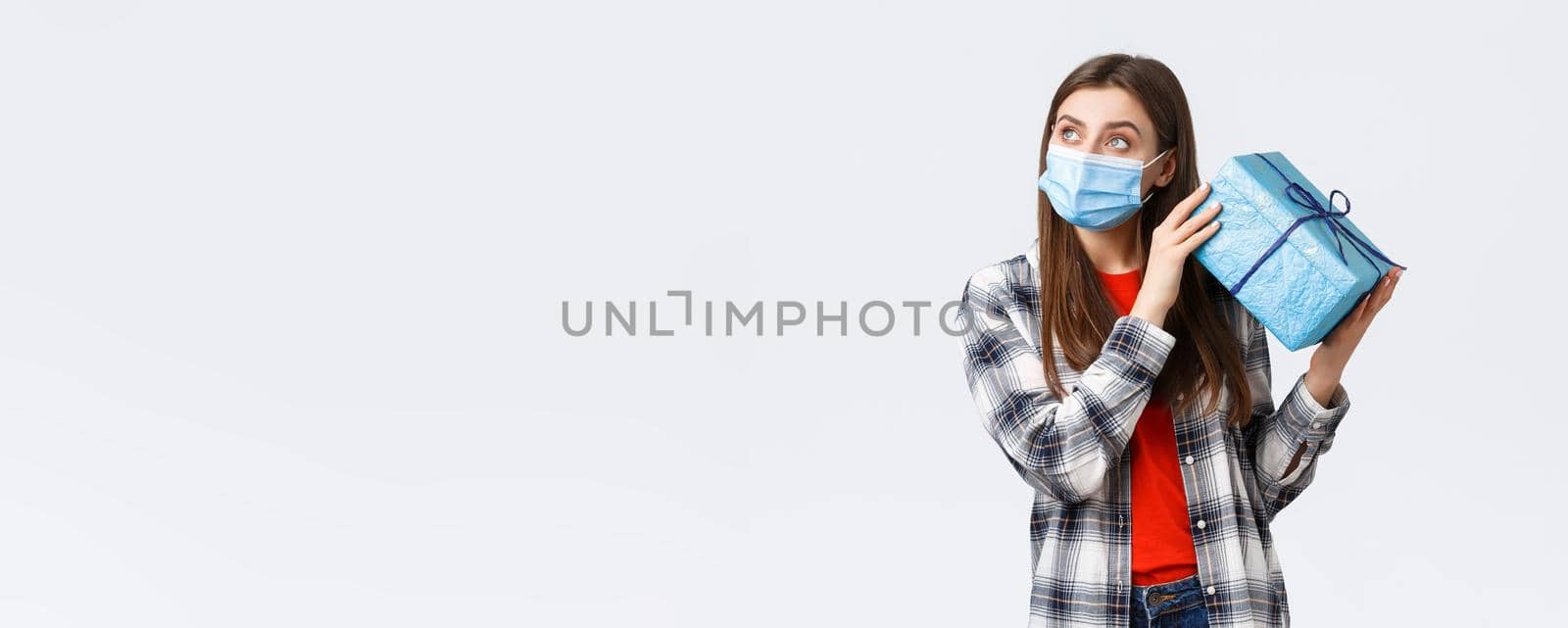Covid-19, lifestyle, holidays and celebration concept. Curious young cute girl celebrating birthday, shaking gift box to guess what inside, look away thoughtful, lean ear to present, white background.