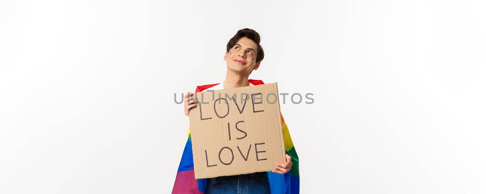 Dreamy young queer person smiling and looking at upper left corner, holding love is love sign for pride parade, wearing Rainbow flag, white background by Benzoix