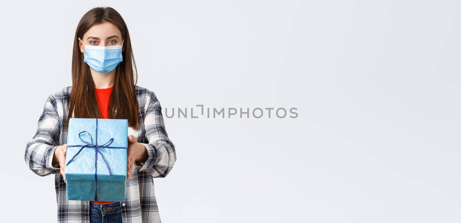 Covid-19, lifestyle, holidays and celebration concept. Cheerful young girl in medical mask stretch hand, handing birthday gift to you, smiling, congratulate with b-day, white background.