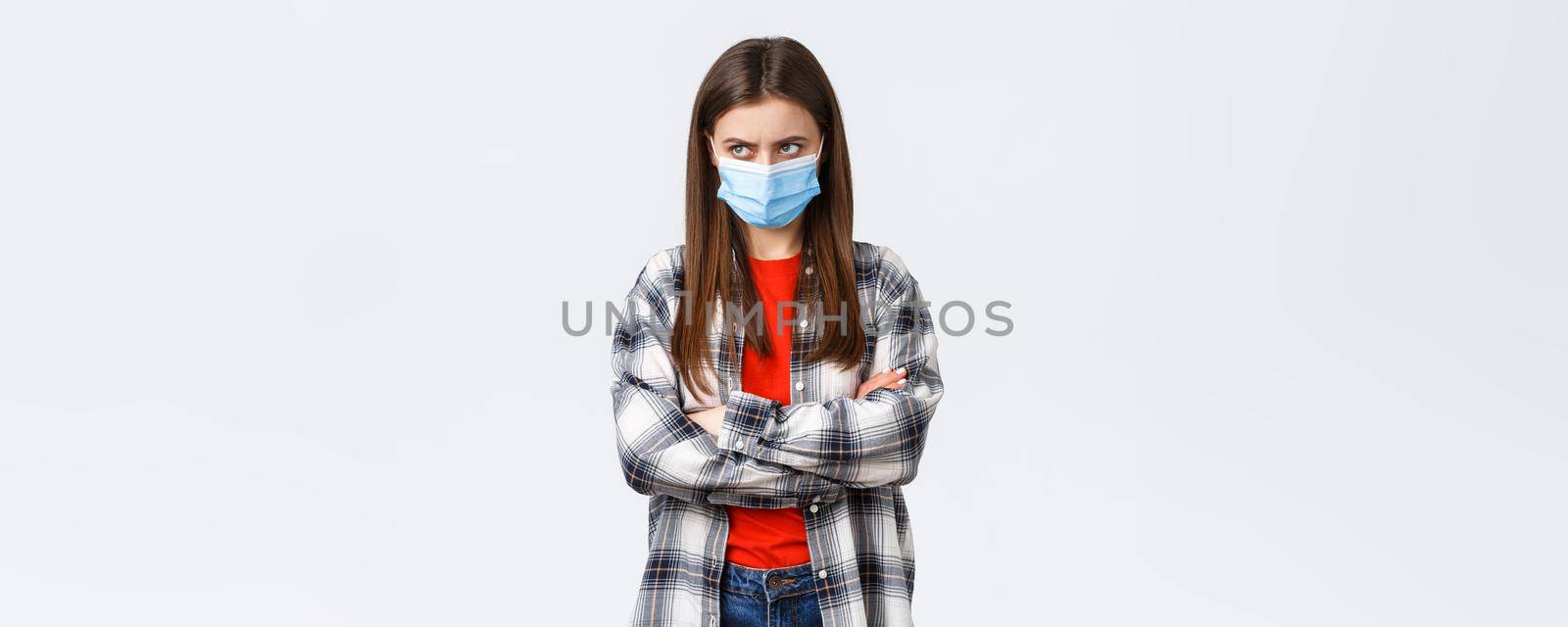 Coronavirus outbreak, leisure quarantine, social distancing and emotions concept. Mad young cute girl in medical mask, cross arms chest, look from under forehead upper left corner, jealous or angry.