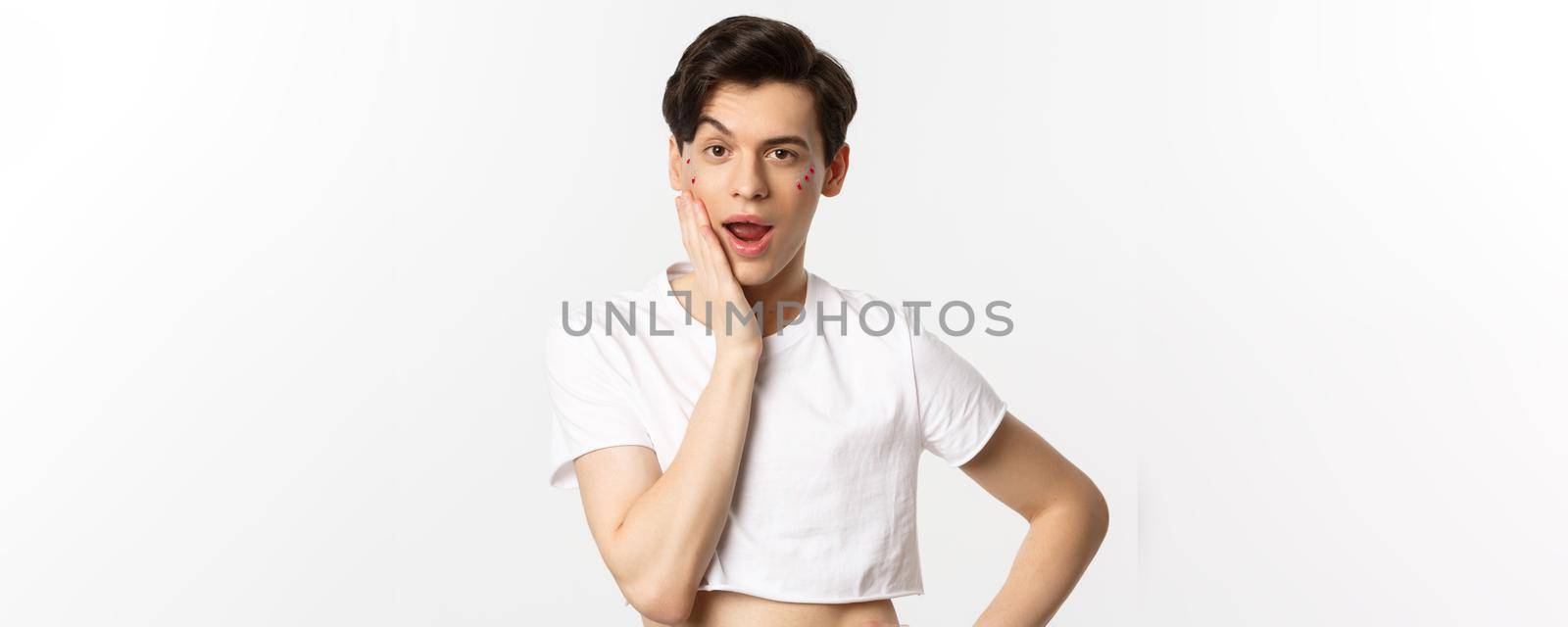 People, lgbtq and beauty concept. Sassy queer person with glitter on face looking cheeky at camera, touching face with hand, standing over white background.