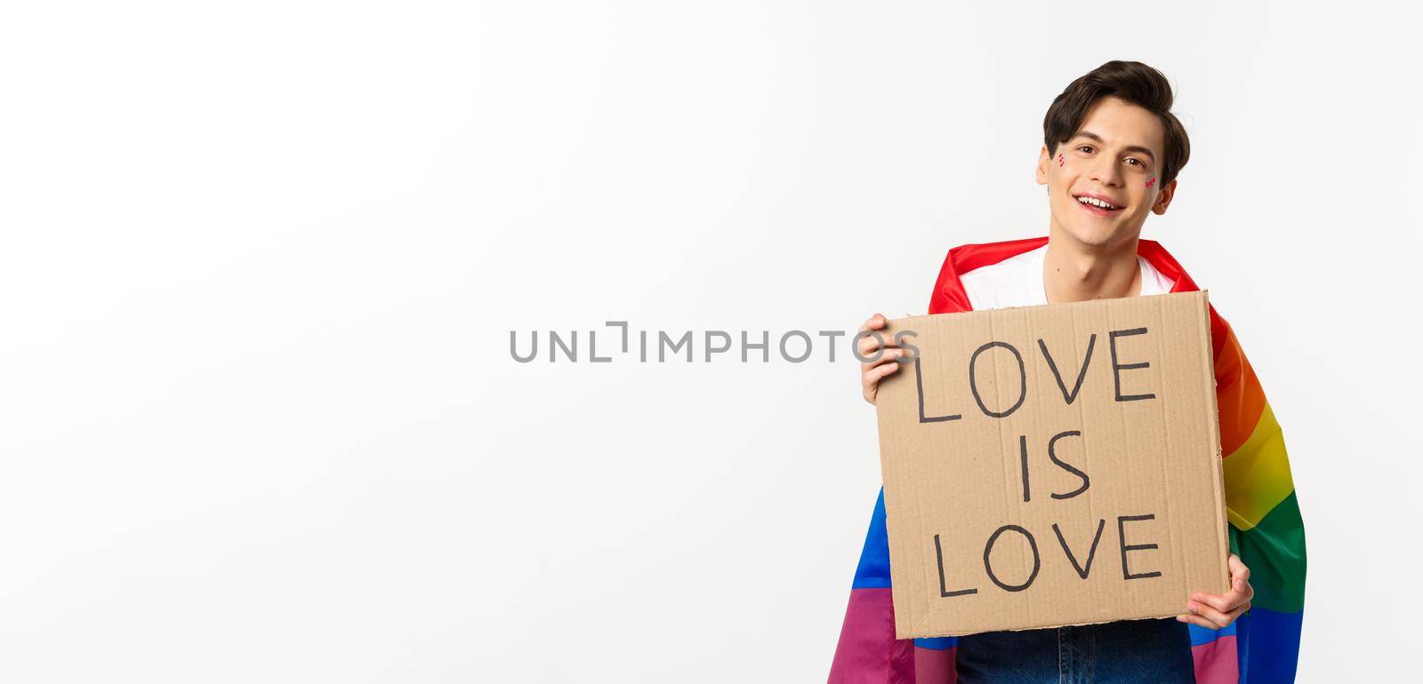Smiling gay man activist holding sign love is love for lgbt pride parade, wearing Rainbow flag, standing over white background.