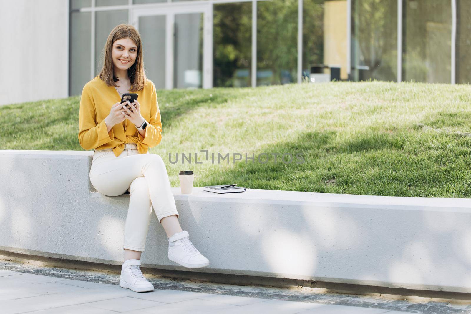 Attractive woman texting message on smartphone and spending time outdoors. Thoughtful lady using cellphone. Smiling woman chatting online on mobile phone near modern buildings