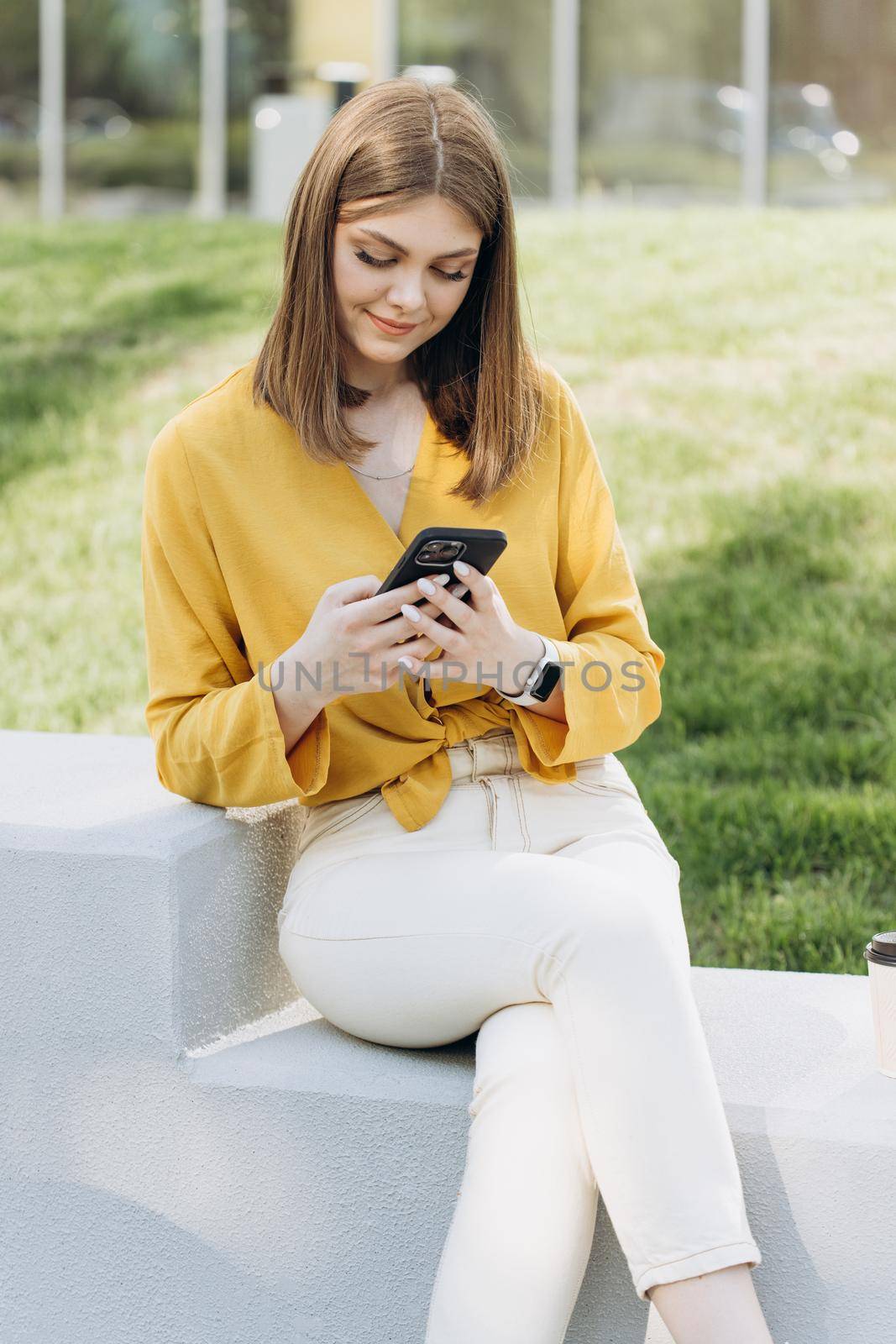 Businesswoman browsing internet on phone, drinking coffee, spending time outdoors. Business woman checking phone content online outdoor. Smiling female freelancer looking phone at remote office by uflypro