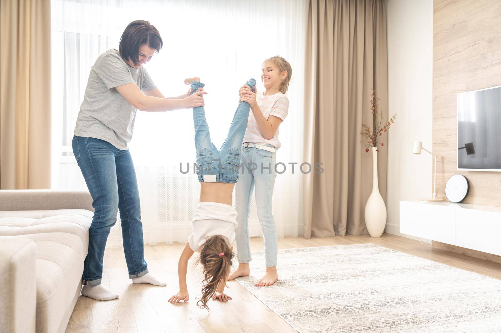 Happy mother and two daughters having fun in living room. mom holding younger daughter upside down by Mariakray