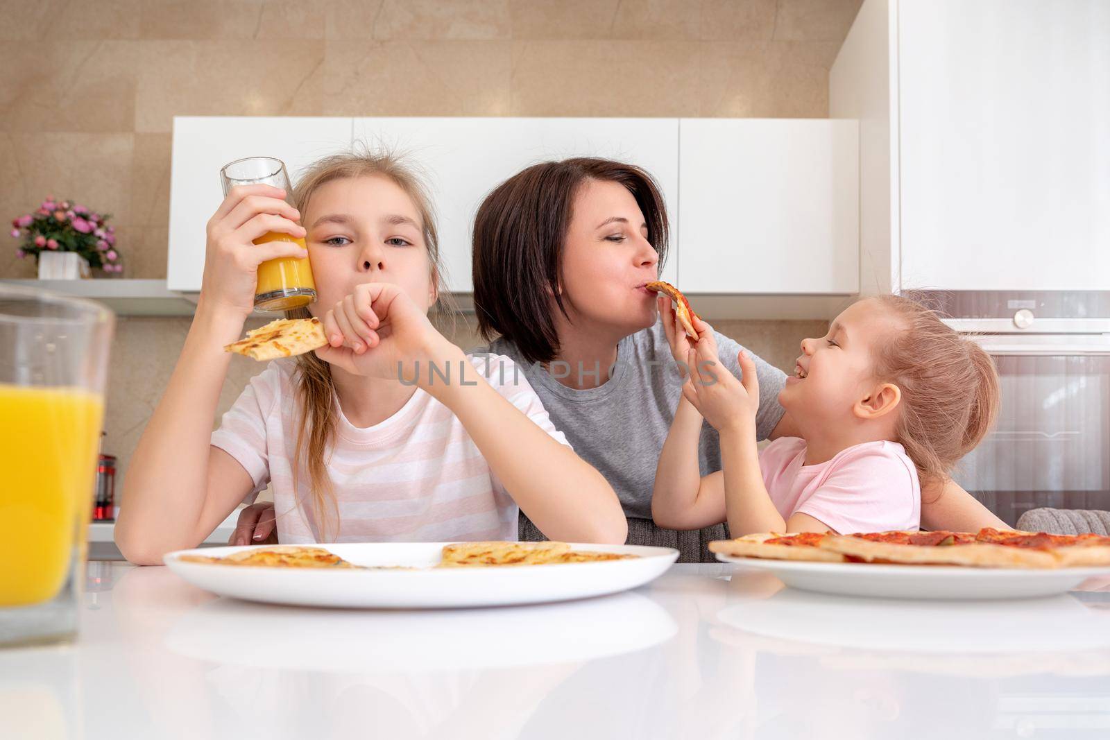 Mother and two daughters eating homemade pizza at a table in kitchen, happy family concept by Mariakray