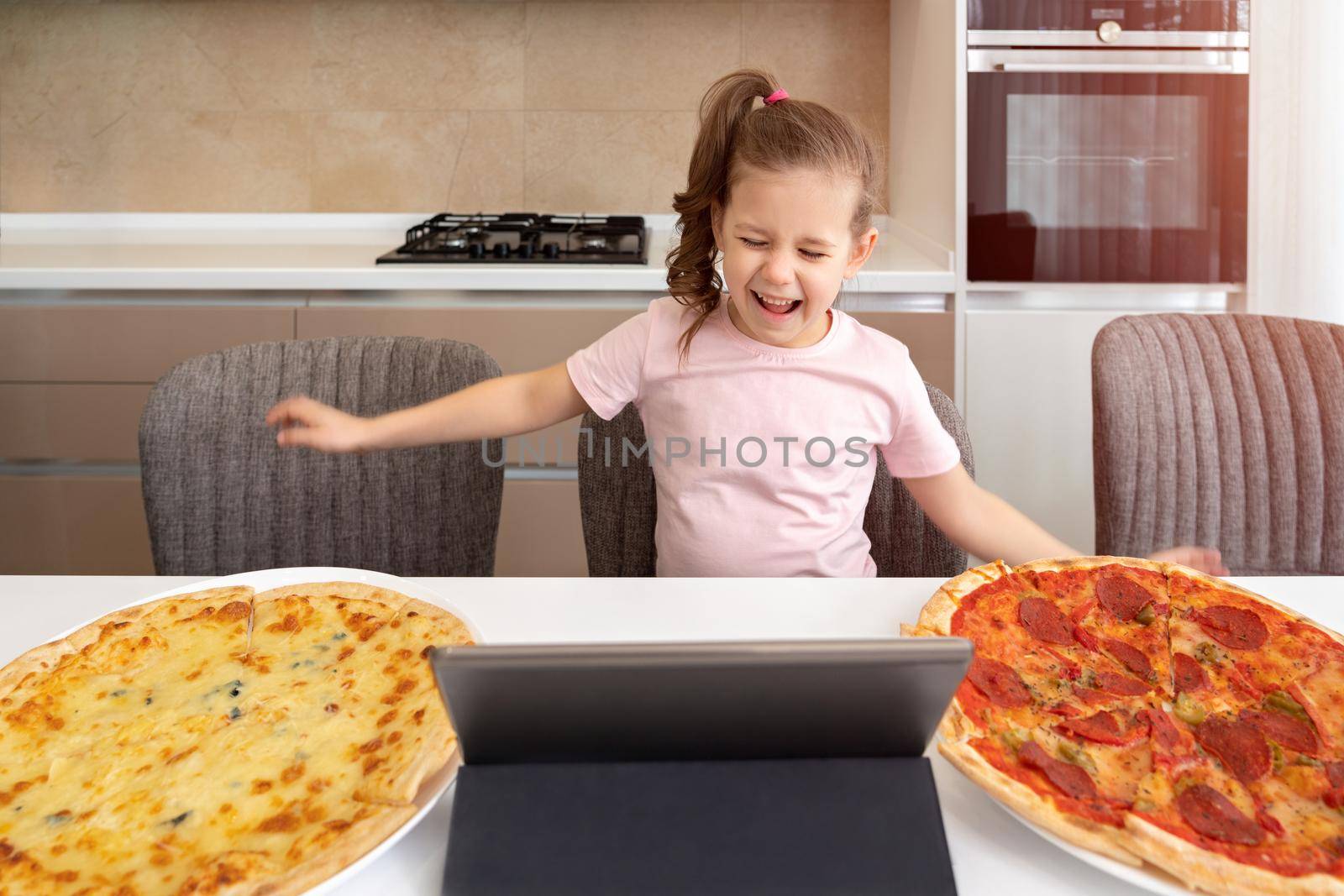 A little girl sitting at a table with a plate of two pizzas and tablet in kitchen by Mariakray