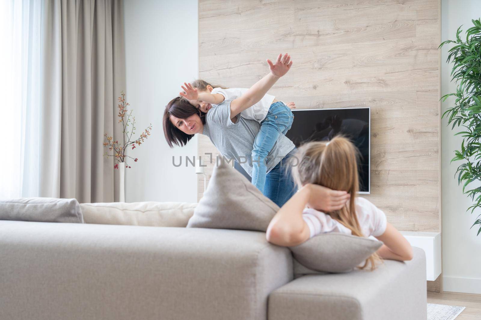 Preteen sad girl sitting on couch while mother having fun with younger sister, jealousy concept by Mariakray