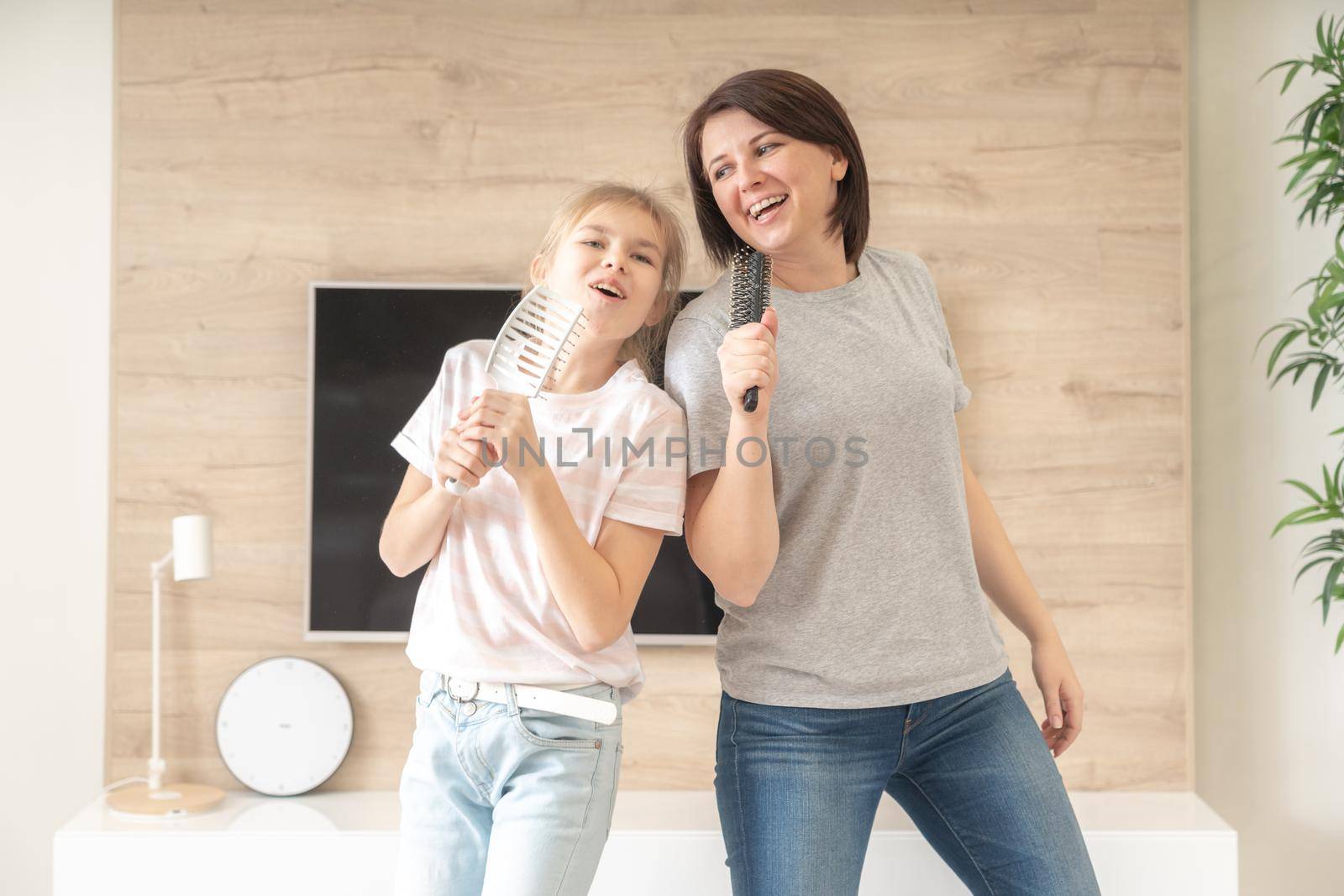 Happy family young adult mother and cute teen daughter having fun singing karaoke song in hairbrushes. mother laughing enjoying funny lifestyle activity with teenage girl at home together. by Mariakray