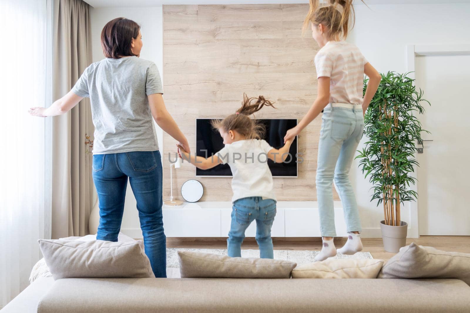 Mother and two daughters having fun jumping on a sofa in living room by Mariakray