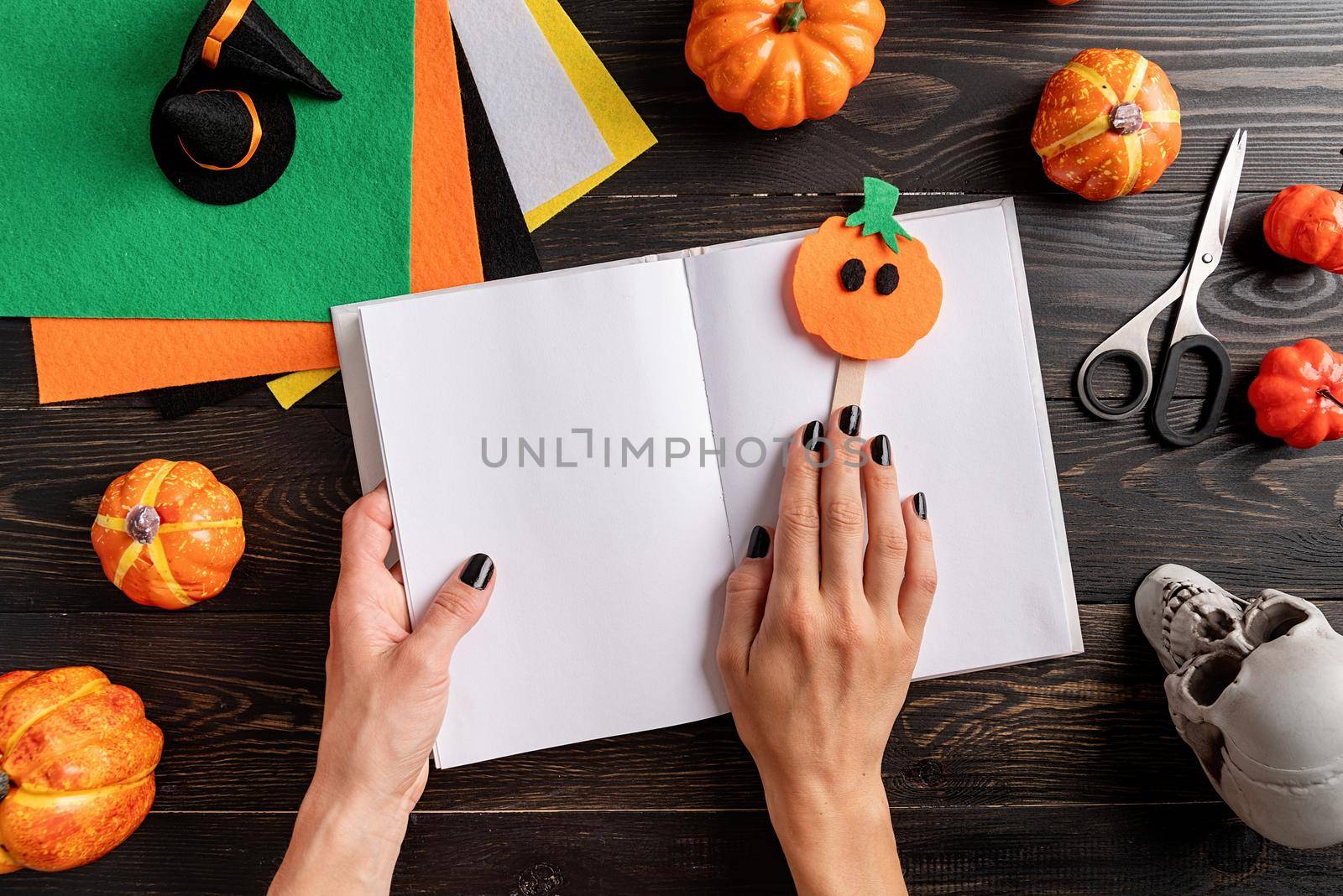 Happy Halloween concept. Making Halloween pumpkin bookmark craft. Woman hands with black nails holding pupmkin bookmark in a blank opened book.