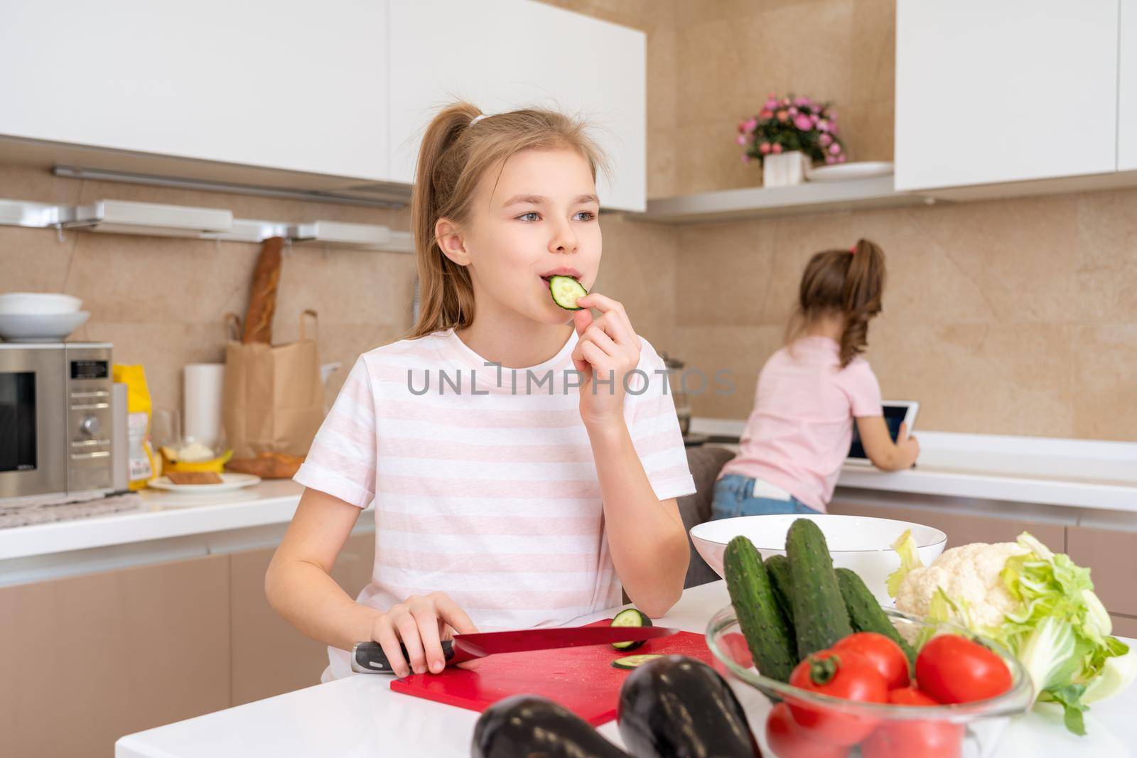 Preteen girl cutting and eating cucumber with knife on a table in kitchen by Mariakray