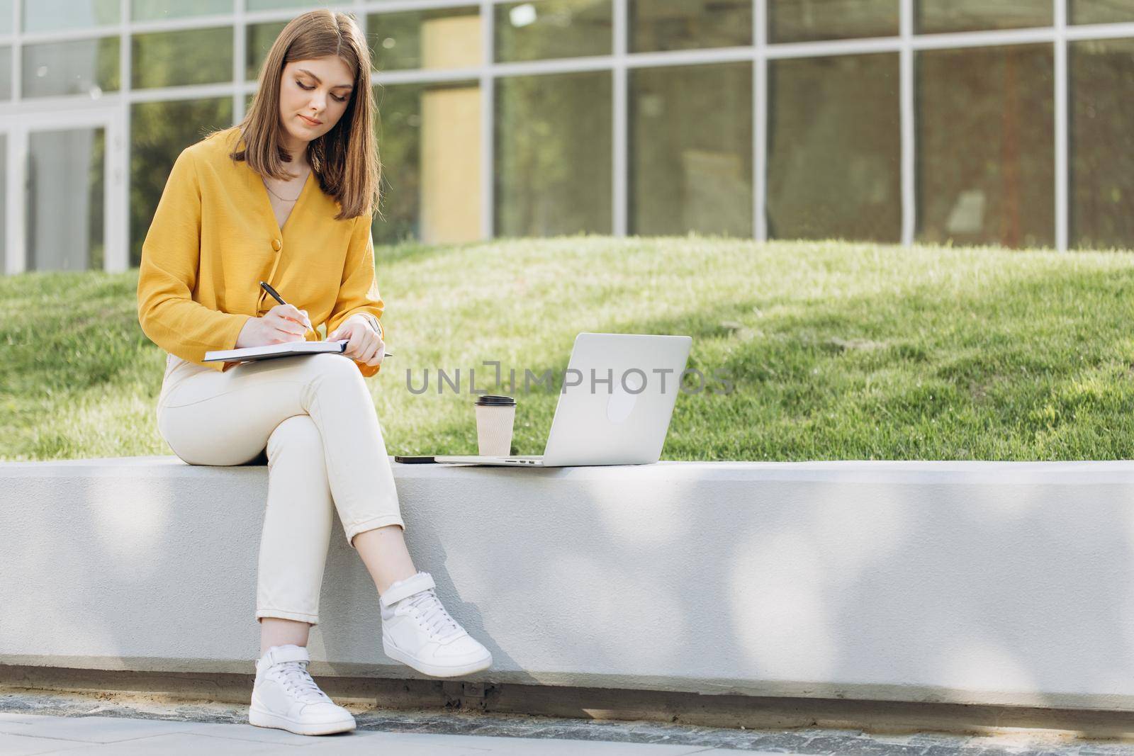 Student girl watching lesson online and studying outside home. Young woman taking notes while looking at computer screen following professor on video call. Girl student studying sitting outside by uflypro