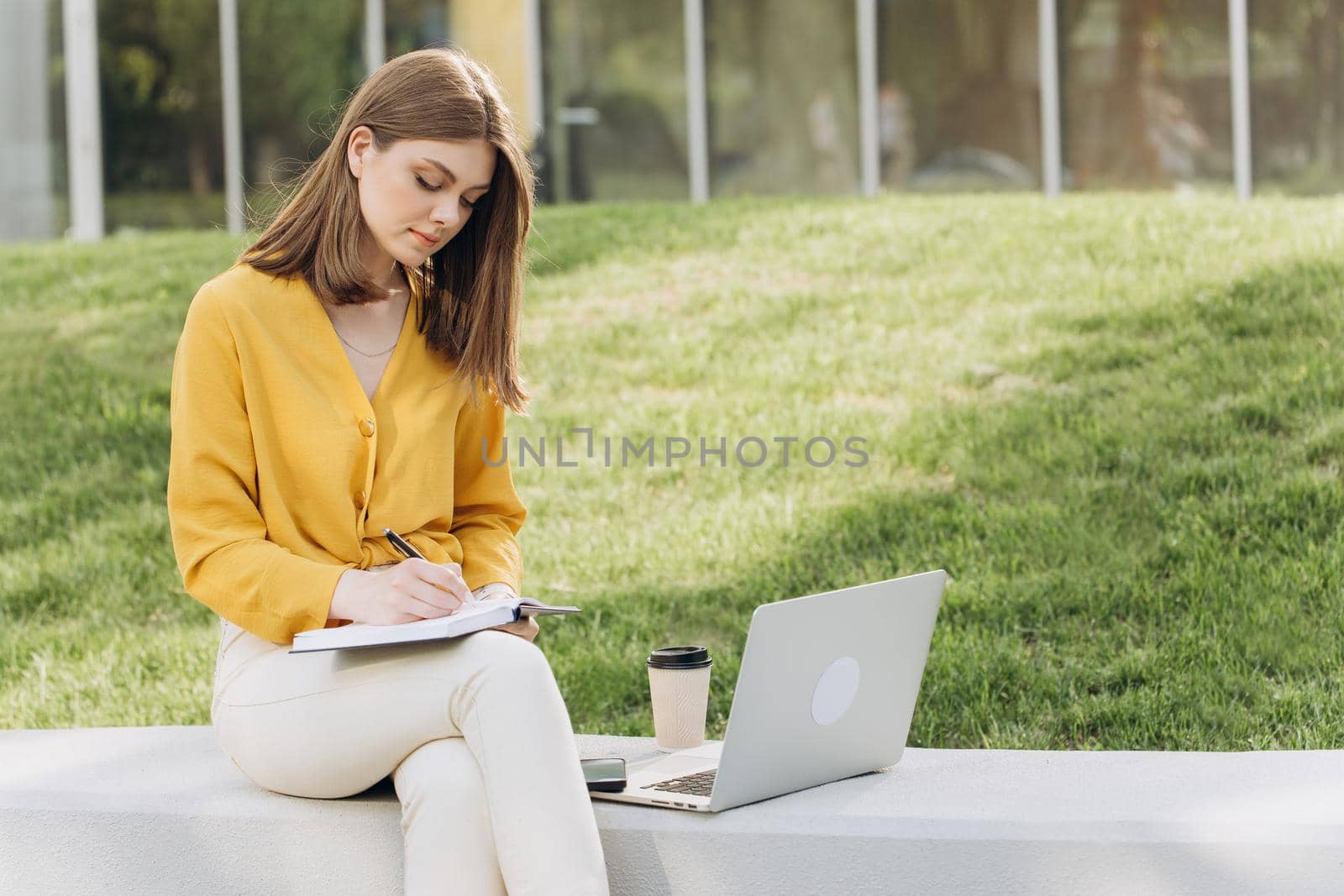 Young caucasian woman university student learning online using laptop computer, taking notes, watching webinar or virtual education remote class studying outdoor sitting outside uni campus area.