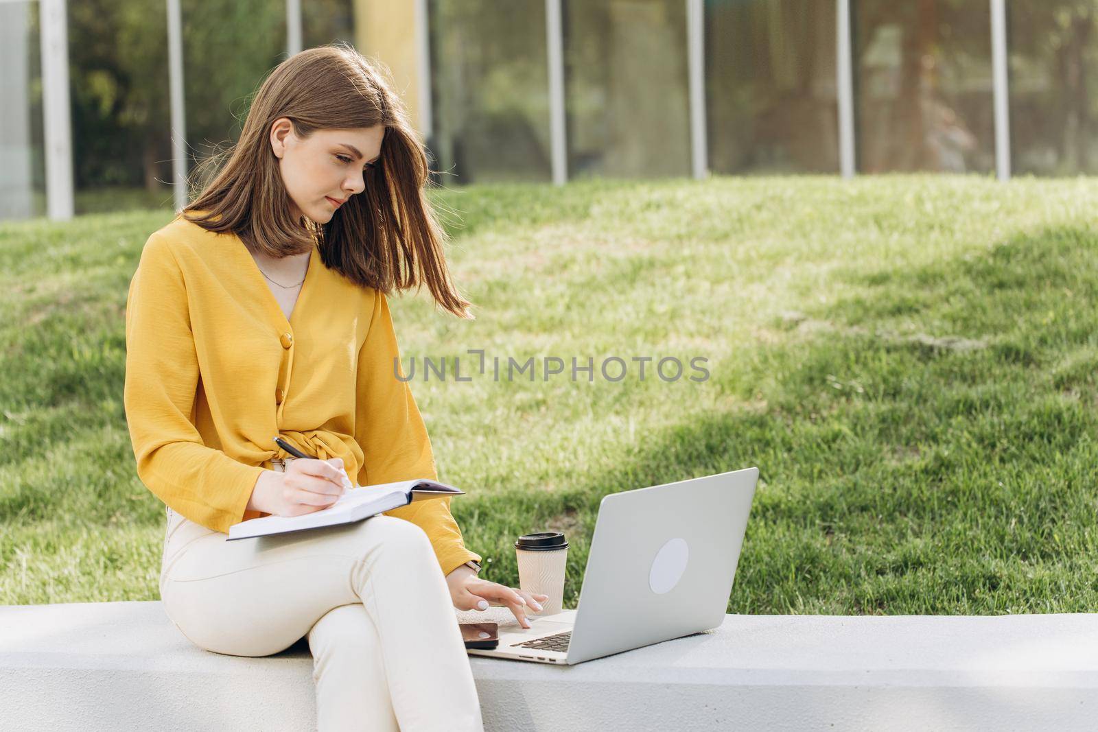 Portrait of focused student girl doing homework next to laptop computer. Teenage girl writing in notebook in front of laptop outdoors. Pretty girl studying at home education by uflypro