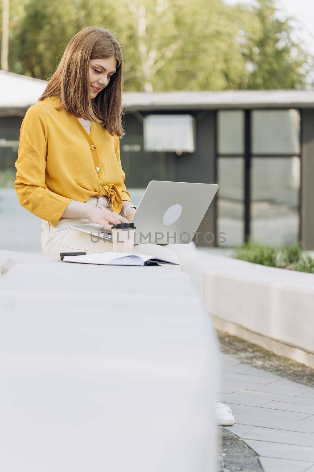 Young european business woman freelancer sitting on bench working with laptop in city park on modern urban street background. Busy worker freelancer working on modern tech notebook device. by uflypro
