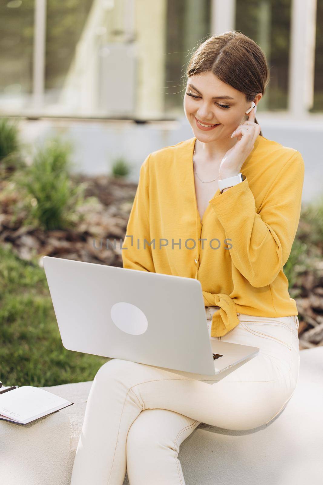 Happy european girl enjoying break time and soft dance. Portrait of beautiful young caucasian woman listening to favorite music in wireless headphones while sitting on bench using laptop computer.