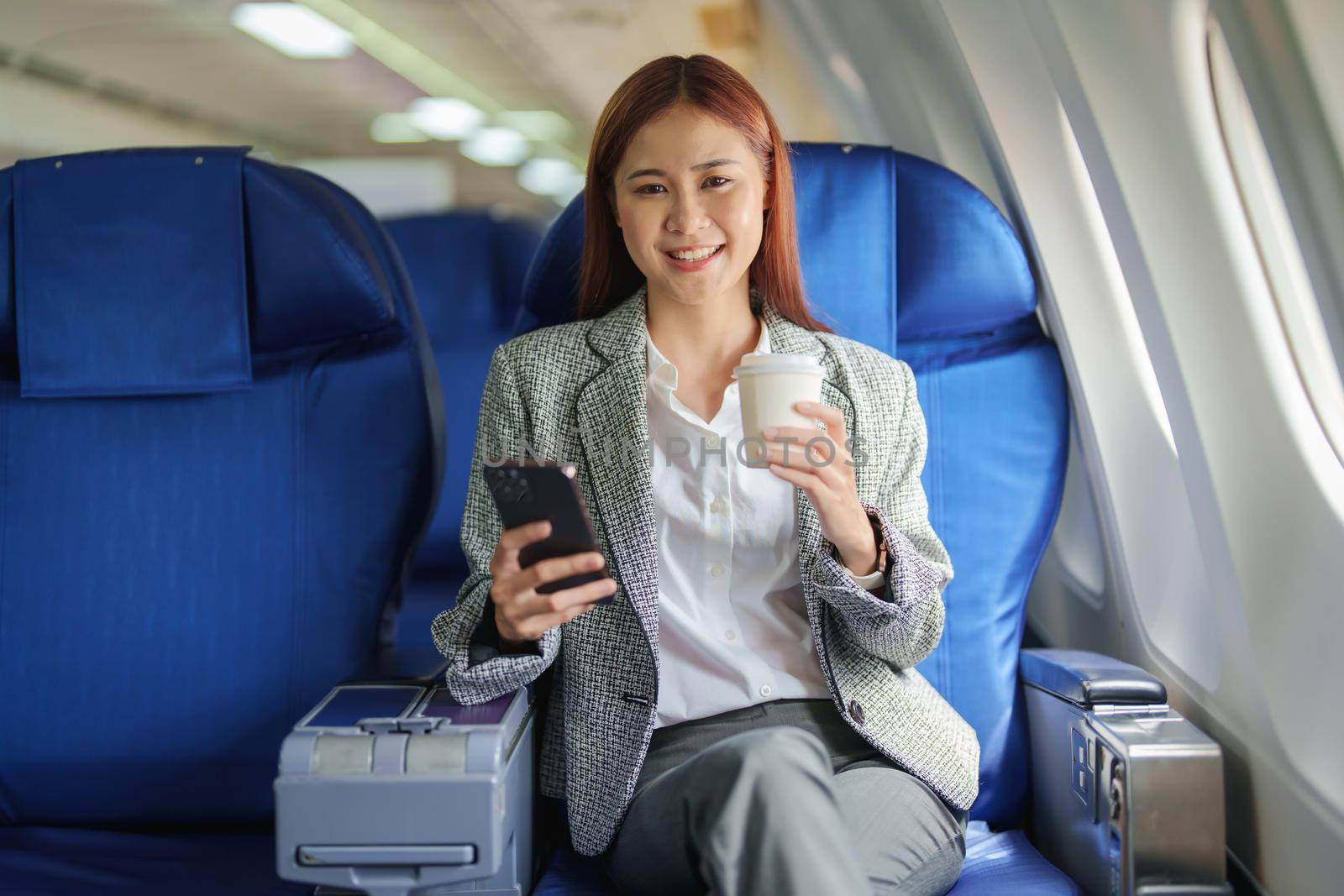 portrait of A successful asian business woman or female entrepreneur in formal suit in a plane sits in a business class seat and uses a smartphone with drink coffee during flight, relax concept by Manastrong
