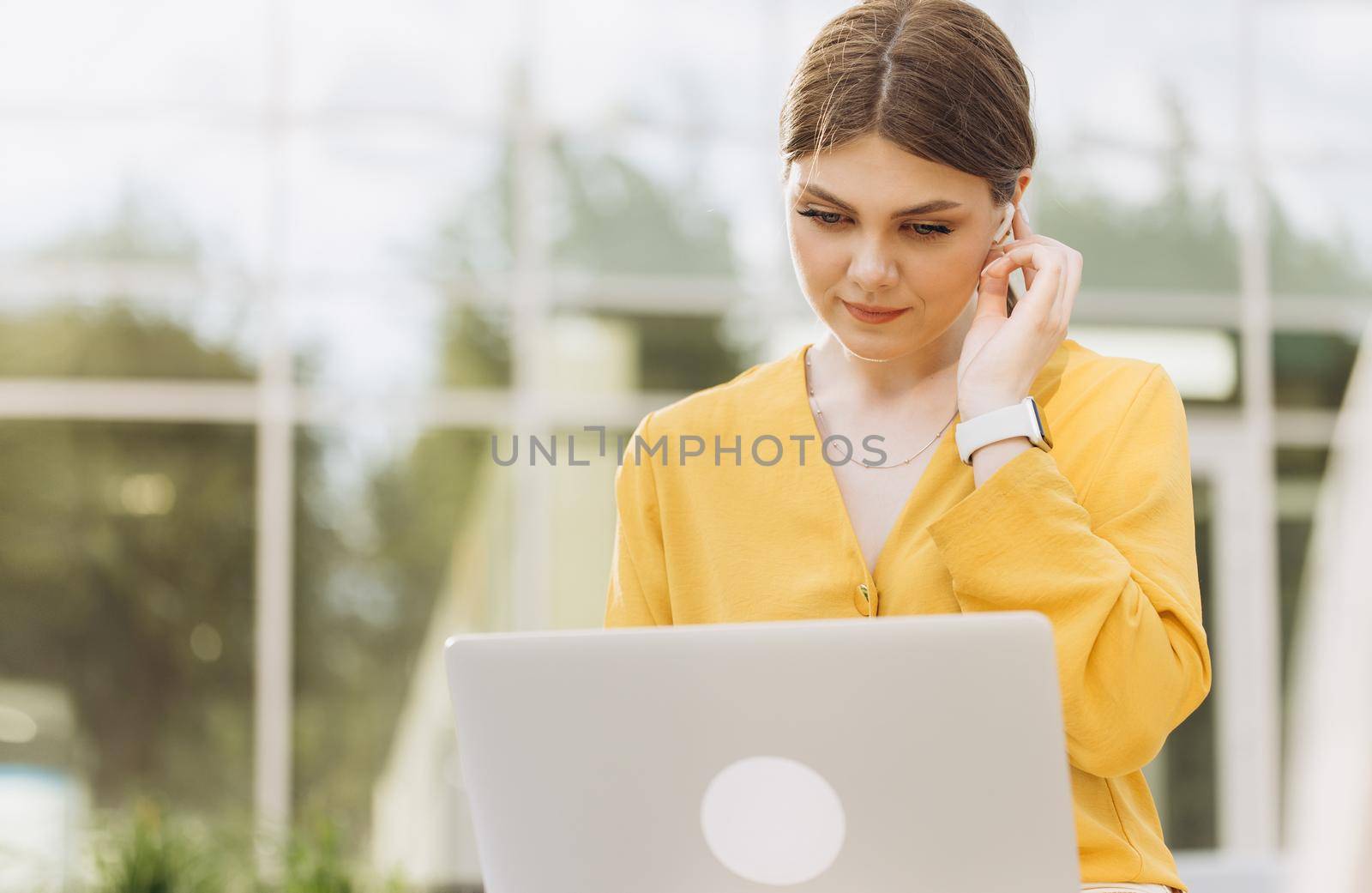Focused businesswoman using wireless earphones working on laptop computer near modern office. Female manager typing on laptop keyboard. Portrait of smiling business woman looking at laptop screen.