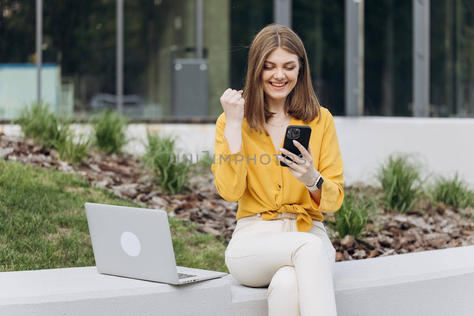 Surprised lady celebrating victory on smartphone outdoor. Portrait of happy business caucasian woman enjoy success on mobile phone. Joyful girl reading good news on phone.