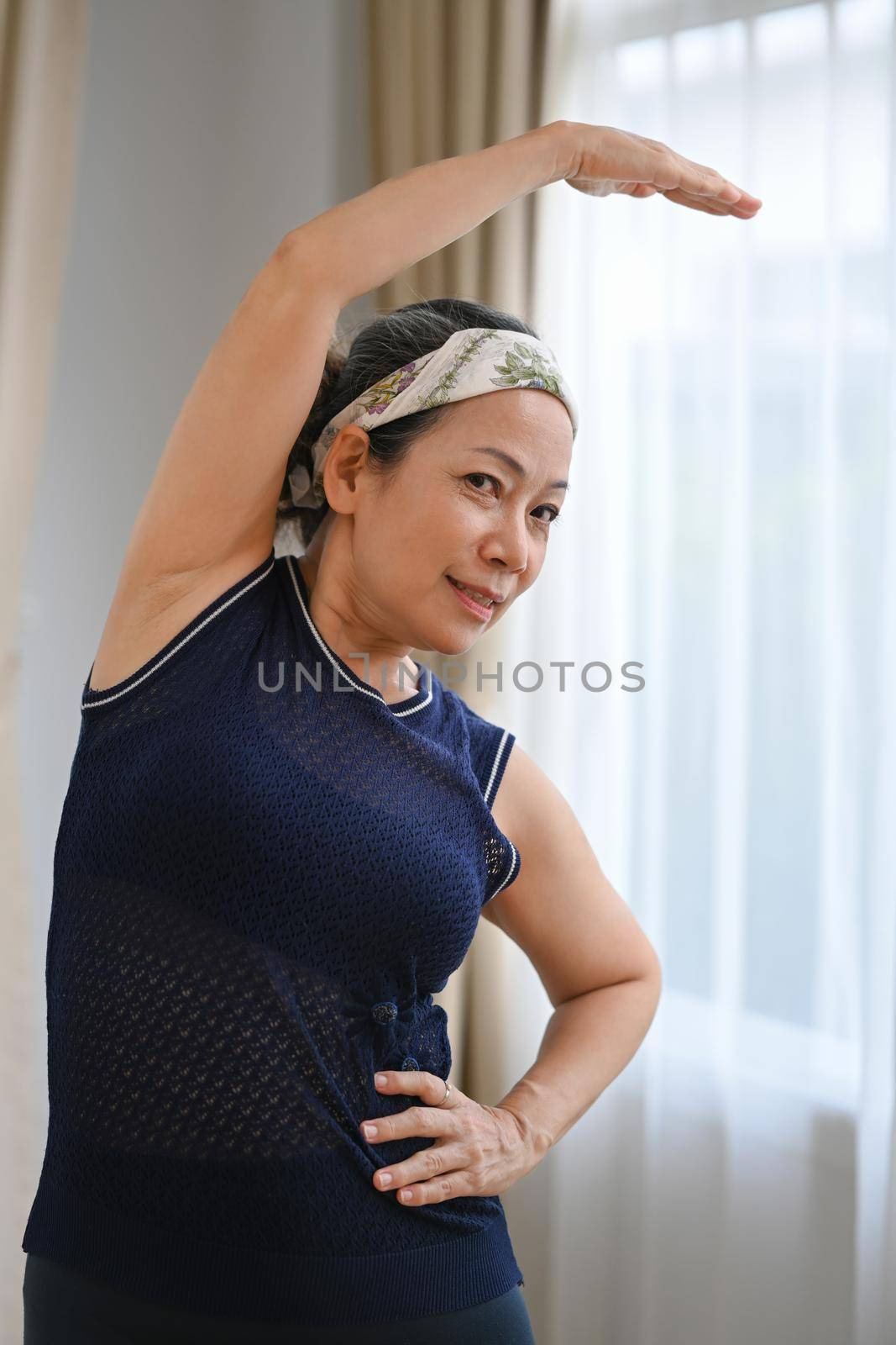 Happy middle aged woman doing her workout routine, exercising indoors, stretching muscles. Health lifestyle concept by prathanchorruangsak