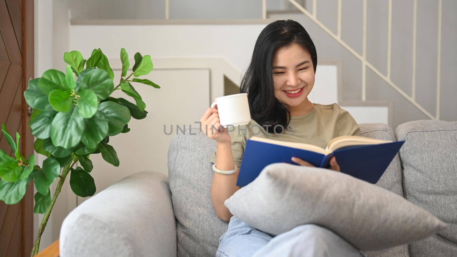 Relaxed young woman reading book and drinking hot coffee on comfortable sofa at home by prathanchorruangsak