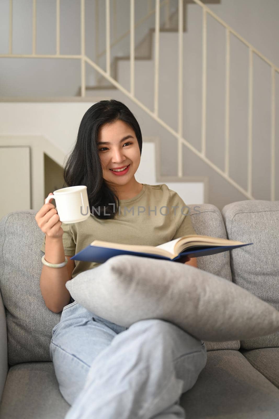 Smiling young woman drinking coffee and reading book on couch, spending leisure weekend at home.